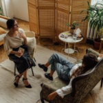 Two women engaged in a psychotherapy session in a warm, inviting interior with plants and natural lighting.