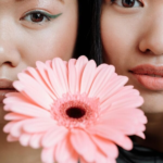 Close-up portrait of two women holding a pink gerbera in front of their faces, with vibrant makeup.