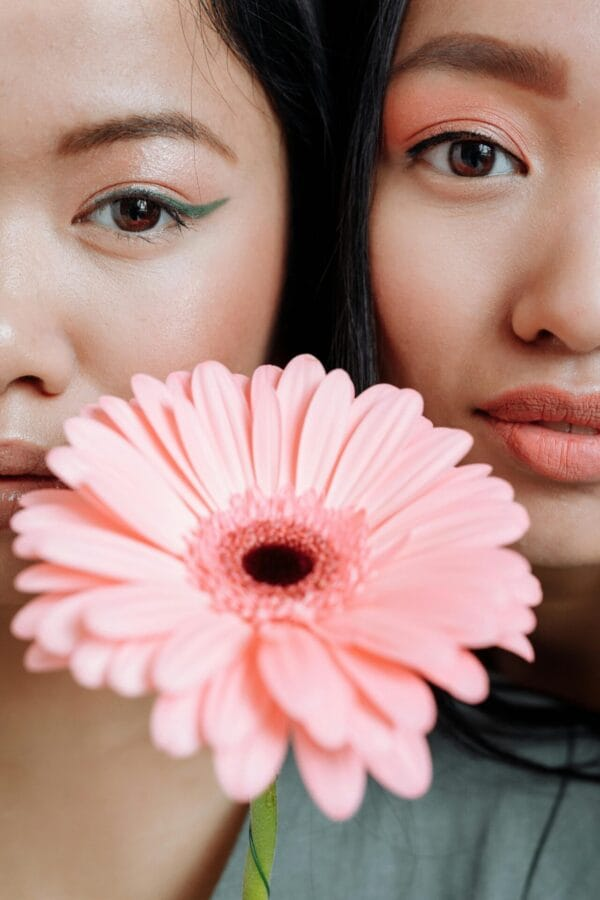 Close-up portrait of two women holding a pink gerbera in front of their faces, with vibrant makeup.