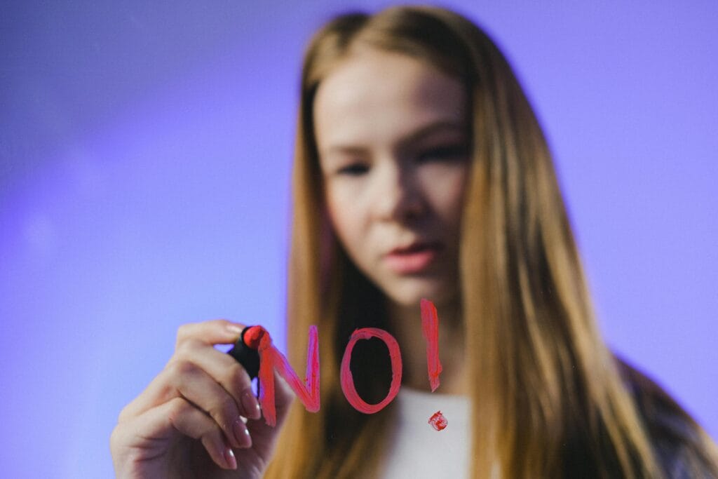 Close-up of a teenage girl writing 'NO!' on glass with a red marker against a blue background.