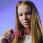 Close-up of a teenage girl writing 'NO!' on glass with a red marker against a blue background.