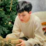 A teenage boy in a sweater holds a wrapped gift by the Christmas tree.