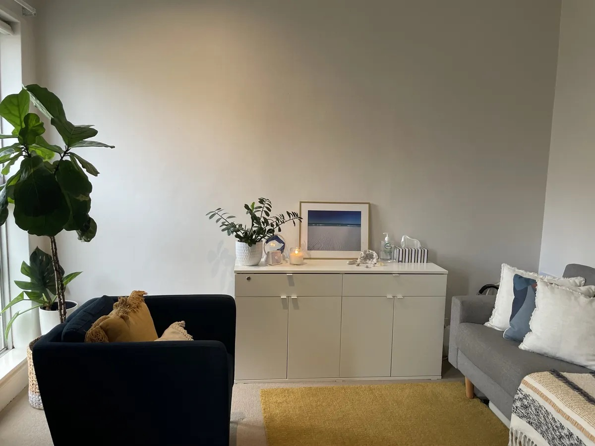 A light, calm therapy room with a navy armchair, a grey couch with cushions, a yellow rug and indoor plants