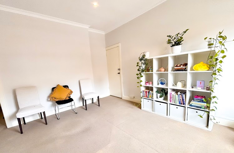 A bright, quiet waiting room with three chairs, a cushion and a white bookshelf with plants, books and toys